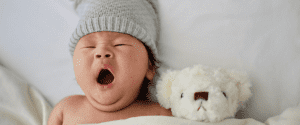 Infant in bed wearing a gray skull cap beanie next to small teddy bear yawning